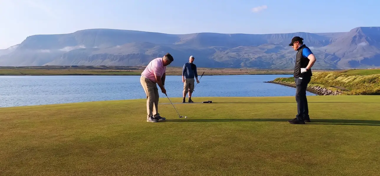 Three golfers on a putting green beside the water with mountains in the background