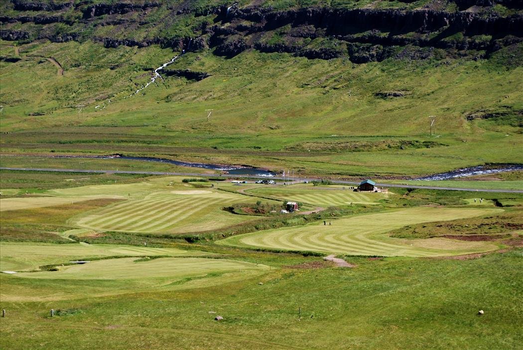 Aerial view of the fairways and clubhouse at Ekkjufellsv&ouml;llur, with a waterfall on the mountainside behind